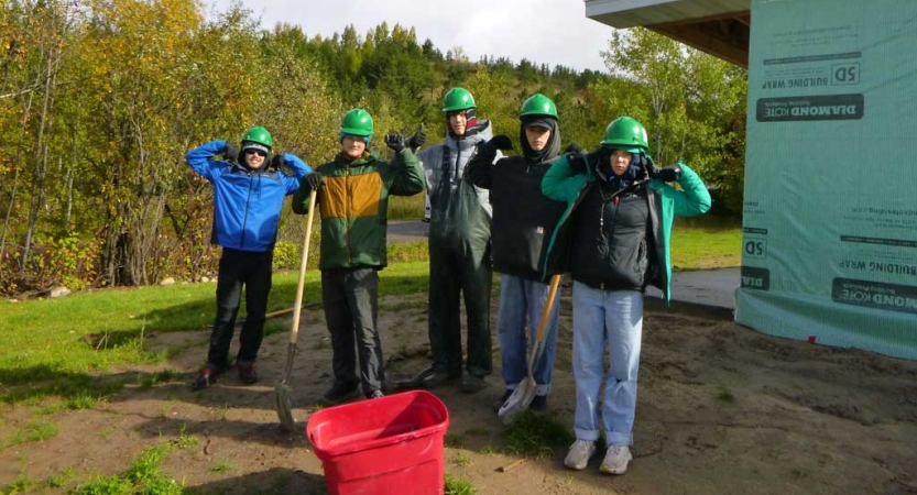 Five students wearing green helmets hold tools near a building site. 